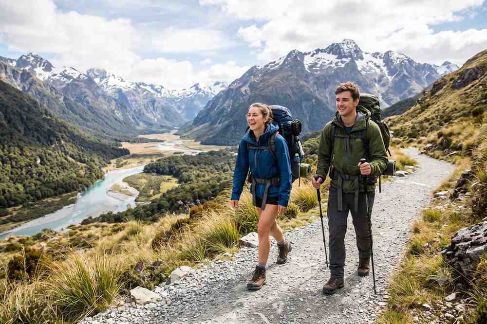 Two happy Kiwis with backpacks and trekking poles hiking an alpine trail, having successfully managed their booking for premier hiking trails in New Zealand.